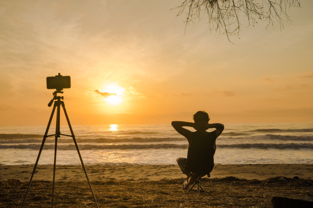 剪影电话三脚架和男人坐在沙滩椅子上录制视频和照片海滩与日出和大海的背景图片下载