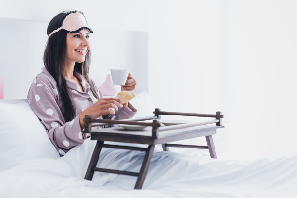 可爱甜美的年轻女子睡衣微笑着坐在床上吃早餐的照片在室内，家里的房间图片下载