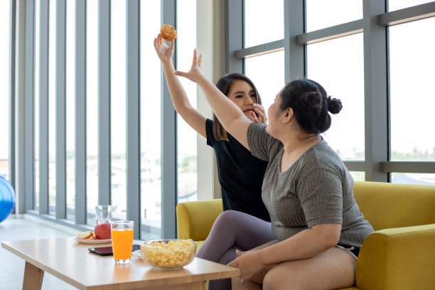 超重的妇女和亚洲女孩喜欢在家里的沙发上吃食物图片下载