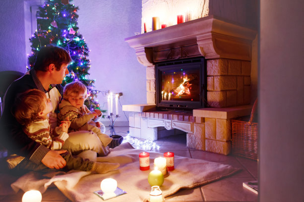 父亲和两个蹒跚学步的小男孩坐在烟囱、蜡烛和壁炉旁，看着炉火。家庭庆祝圣诞节。以圣诞树和灯光为背景。孩子们喜欢礼物图片下载
