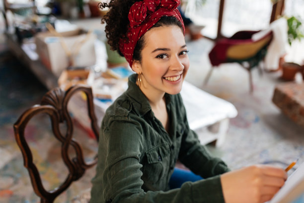 在现代舒适的美术车间里，年轻开朗的深色卷发女子坐在椅子上在画布上画画，快乐地看着镜头图片下载