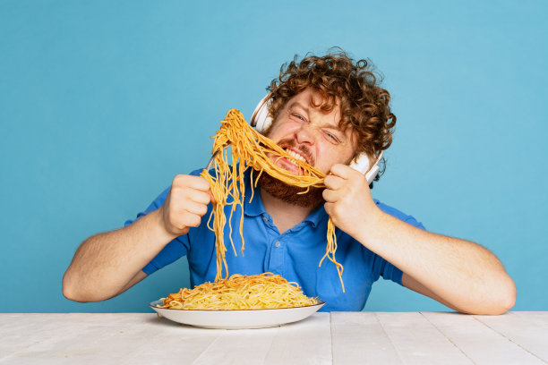 疯狂的年轻红胡子男人品尝着大份的面条，意大利面孤立在蓝色的工作室背景下。图片下载