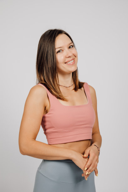 美丽的女人在工作室摆姿势。这是一张快乐的年轻黑发女郎的照片，她穿着米色上衣和灰色瑜伽或健身裤，站在灰色孤立的背景上，微笑着，一只手放在另一只手上图片下载