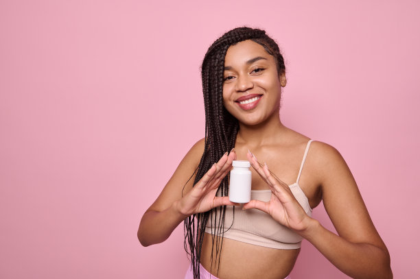 迷人的非洲妇女露出露齿的微笑，拿着一瓶营养补充剂或维生素，为皮肤和头发的美丽和健康，摆姿势在粉红色的背景与广告文字的空间。图片下载