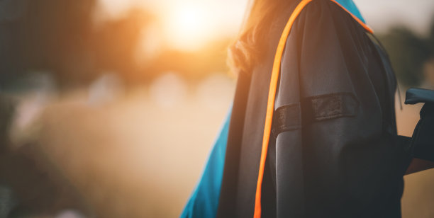 毕业的学生，头戴黄流苏的黑帽子，手拿毕业证站在夕阳下，祝贺概念教育。毕业典礼，祝贺大学毕业生。图片下载