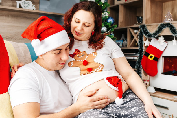 幸福的年轻家庭，年轻人坐在椅子上，把怀孕的妻子抱在膝上，用手指着她怀孕的肚子，他们穿着轻便的t恤和圣诞老人的帽子。恭贺新禧，合家欢乐。一个年轻的家庭希望有个孩子。图片下载