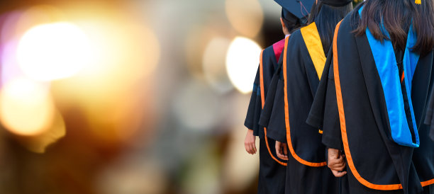 毕业生穿着传统的长袍和流苏帽站在夕阳下，手拿文凭，概念教育祝贺。毕业典礼，祝贺大学毕业生。图片下载