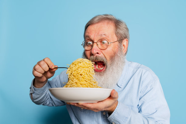 情绪激动的头发灰白的老男人吃着一大块面条，意大利面孤立在蓝色的工作室背景下。图片下载