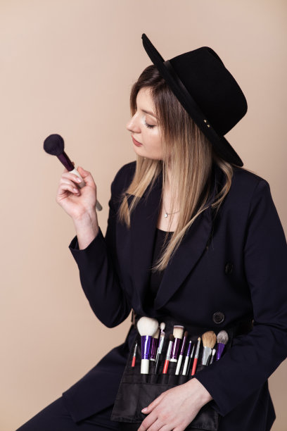年轻可爱的女性设计师与化妆师腰带和画笔。专业的化妆工具。米色工作室背景。图片下载