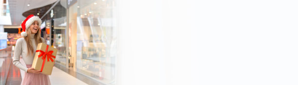 快乐的年轻女子，戴着圣诞帽，拿着礼盒在购物中心。假日购物和礼物。销售。购物中心。副本的空间。横幅图片下载