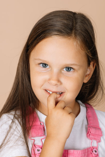 小女孩咬着指甲，闪亮的眼睛微笑着看着镜头，穿着亮粉色的连体衣和白色的t恤，米色的背景图片下载