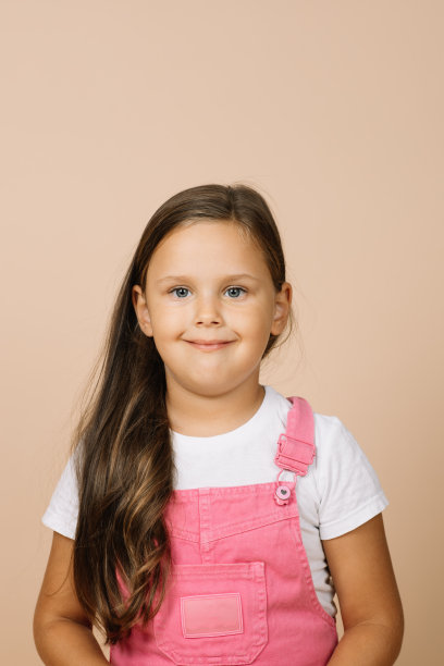 近脸孩子明亮的闪亮的眼睛和平静的快乐的宽微笑看着镜头穿着亮粉色的连体衣和白色的t恤在米色的背景图片下载