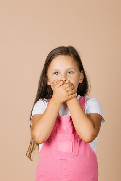照片中，小女孩闭着嘴，不说话，眼睛闪闪发光，穿着亮粉色的连体衣和白色的t恤，米色的背景图片下载