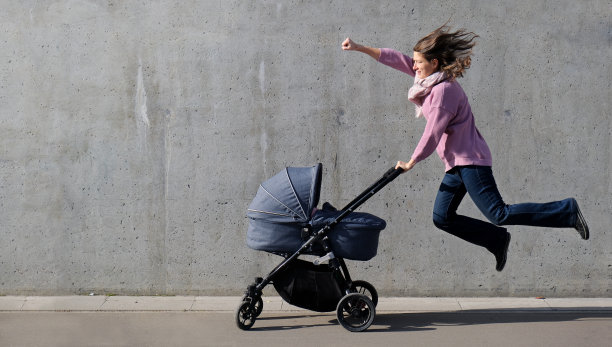 在灰色的混凝土墙背景下，超级英雄母亲带着刚出生的婴儿飞翔。强壮有力的女人和婴儿车看起来像超级英雄。图片下载