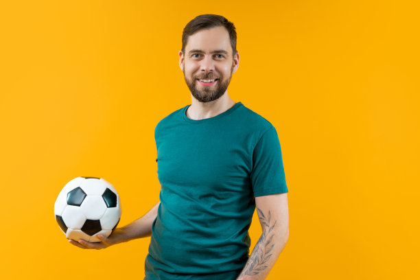 年轻英俊的蓄着胡须的足球支持者的肖像，在黄色背景上孤立地摆姿势，手里拿着一个球图片下载