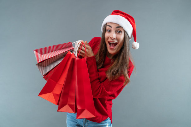 工作室的照片中，激动的漂亮女孩戴着圣诞老人的帽子和红色舒适的针织毛衣，手里拿着一堆购物袋，在灰色的背景下孤立图片下载