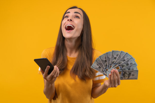 这是一个年轻漂亮的女孩，手里拿着一沓美元钞票，脸上露出惊讶的表情，简直不敢相信自己在智能手机上中了头奖图片下载