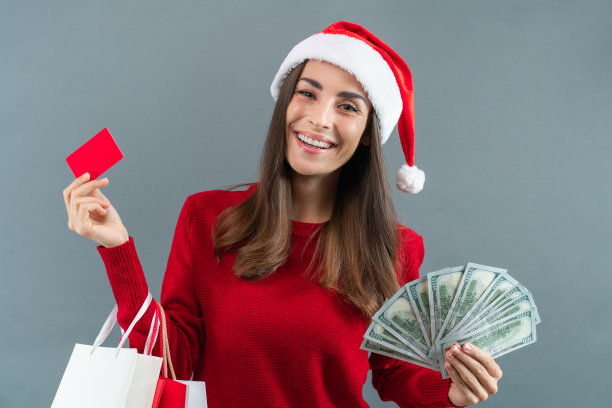 工作室里的一幅照片上，迷人的年轻女子戴着圣诞老人的帽子，手里拿着购物袋、信用卡和一叠美元钞票，背景是灰色的图片下载