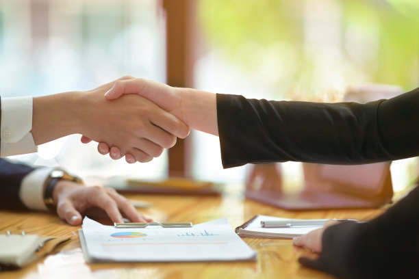 裁剪过的照片显示，商务人士在被文书和办公设备包围的木制办公桌上结束会议或谈判后握手。图片下载