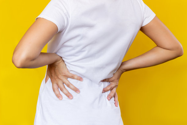 裁剪的照片，一个年轻的女人在白色的t恤，她的下背部与她的手在黄色的背景。背部疼痛图片下载