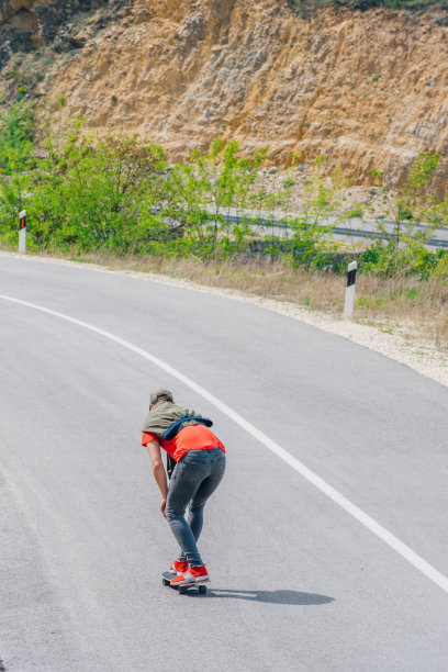 男性白人滑板运动员在一条空路上滑下坡，做一个快速折弯和抓板，同时快速驾驶长板。红t恤，绿帽子，黑牛仔裤。图片下载