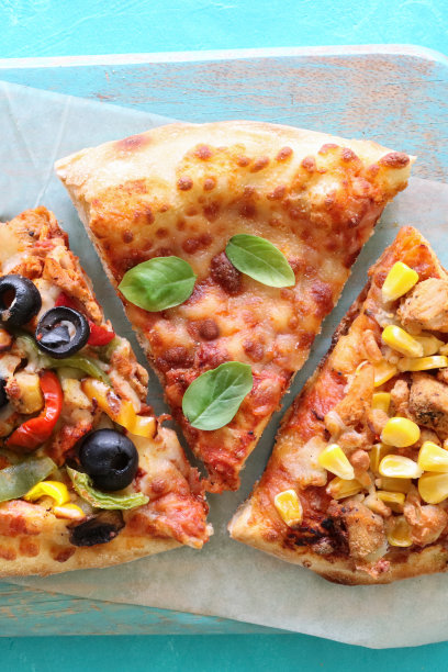 三张带有各种配料的披萨的特写图片，包括鸡肉和甜玉米、玛格丽塔披萨和黑橄榄蔬菜，放在切菜板上，防油纸，蓝色背景，高架视图图片下载