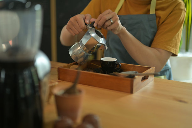 这张照片拍摄的是服务员的手从一个钢制水壶里倒出热咖啡到盛满咖啡豆和豆勺的咖啡杯。图片下载