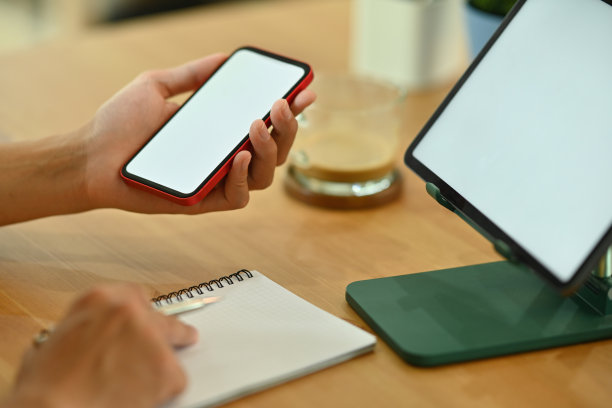 这张照片是一个年轻的自由职业者拿着一个白屏幕的智能手机，在木桌上做笔记，周围是个人设备和平板电脑上的白屏幕数码平板。图片下载
