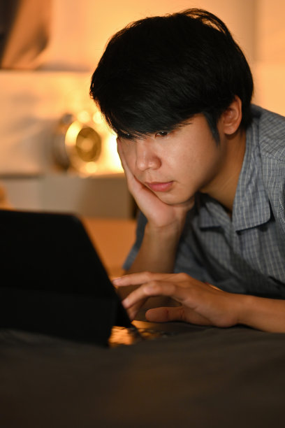 这是一张穿着睡衣的年轻人躺在床上使用数码平板电脑的照片。图片下载