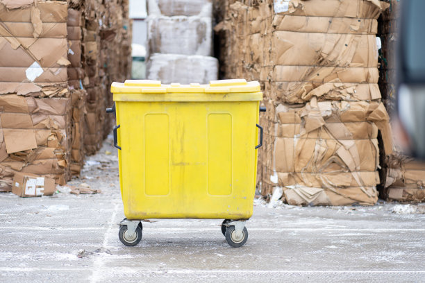 一个黄色的垃圾桶在一个大包装的废纸前的照片。垃圾分类的生产。图片下载