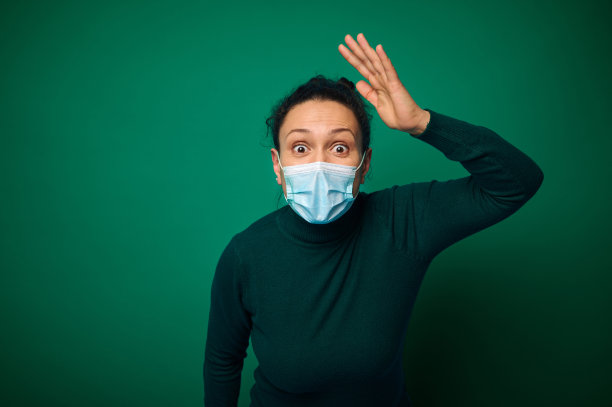 照片中，一名非洲年轻的卷发妇女震惊地瞪大了眼睛，带着惊讶的表情，戴着保护性的医用口罩，绿色的针织毛衣被隔离在彩色的工作室背景上。Omg的概念图片下载