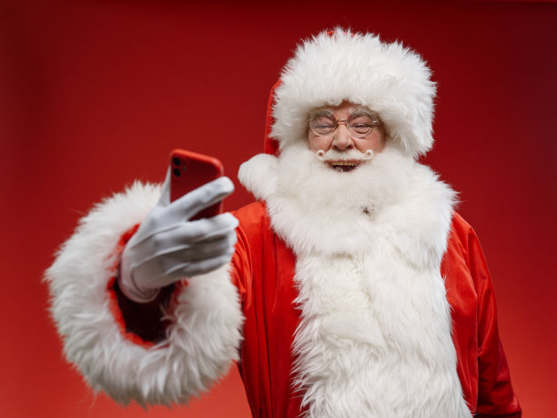 笑着的圣诞老人站在照相馆的亮红色背景上，看着他的红色智能手机图片下载