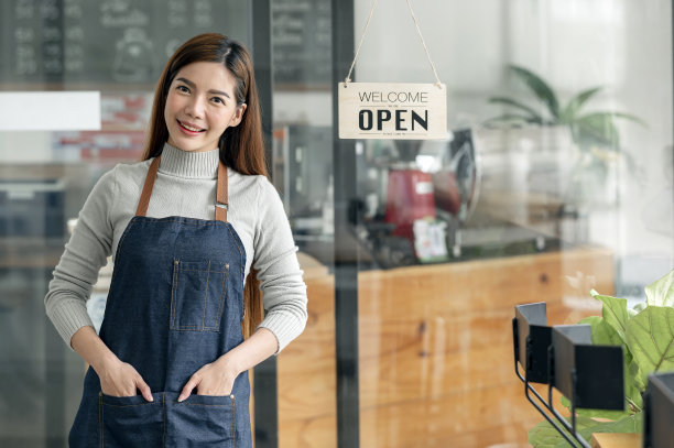 年轻的女企业家，穿着羊毛衫和围裙，站在自助餐厅门口，黑板开着。图片下载