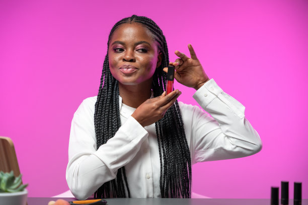 一位年轻迷人的美妆博主穿着白色衬衫，扎着非洲式的辫子，坐在粉红色背景的桌子旁，在镜头前展示着一支口红图片下载