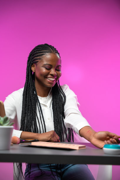 一个年轻迷人的深肤色博主，扎着非洲式发辫，面带微笑地坐在工作室的一张桌子旁图片下载