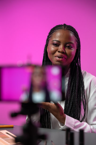 一个年轻迷人的深色皮肤的女博主，扎着非洲式的辫子，身穿白衬衫，坐在工作室的桌子旁，以亮粉色为背景，为她的频道录制新一集图片下载