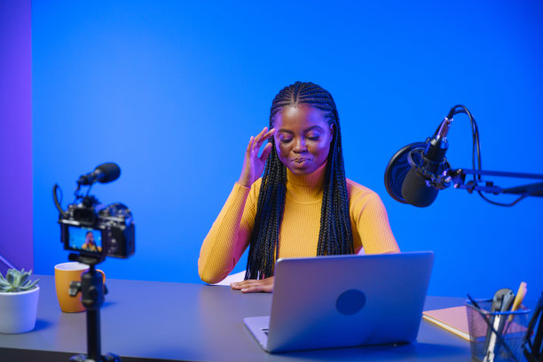 一位年轻的深肤色视频博主，身穿黄色高领毛衣，扎着非洲辫子，正坐在专业工作室蓝色背景的桌子旁，为她的频道录制视频图片下载