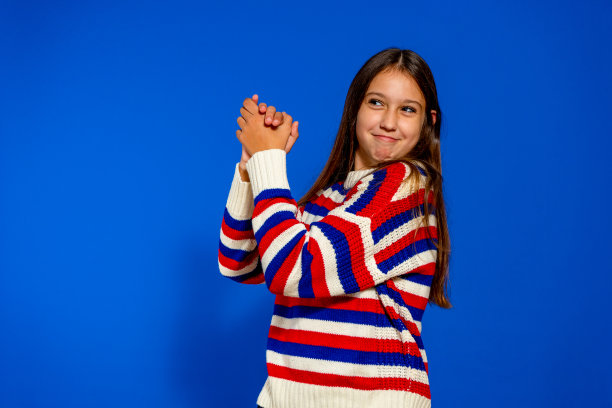 穿着条纹毛衣的白种人女孩在蓝色的墙壁上高兴地鼓掌庆祝，骄傲地微笑着手在一起。图片下载