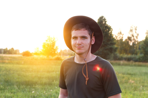 年轻的农民戴着牛仔帽在夕阳下的农田。戴着帽子的千禧一代男子的肖像，站在草地背景上，户外的草地上。农业的概念。本空间图片下载