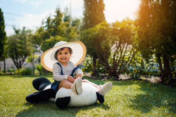 快乐的小女孩戴着一顶特大的白色帽子，正在草地上玩她的玩具熊猫熊图片下载