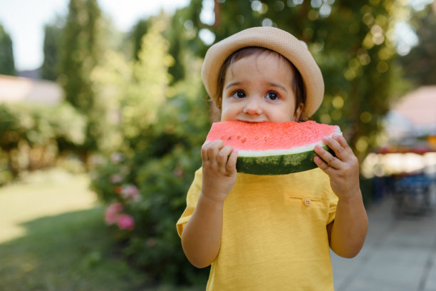 穿着黄色t恤和帽子的2岁小女孩正在吃一片非常有趣的西瓜。图片下载