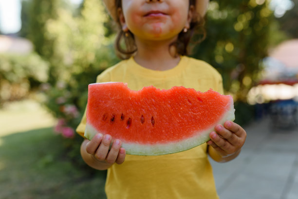 2岁的小女孩穿着黄色t恤，戴着帽子，手里拿着一片西瓜。图片下载