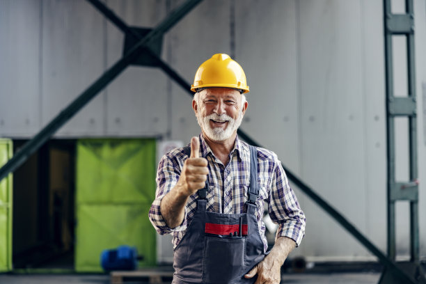 工厂生意很好。一位头戴安全帽、面带微笑的老员工站在工厂里，竖起大拇指。图片下载