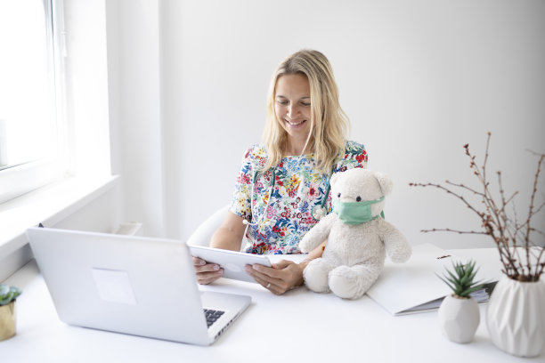 漂亮的金发小儿科医生拿着听诊器坐在笔记本前，手里拿着白色的泰迪熊图片下载