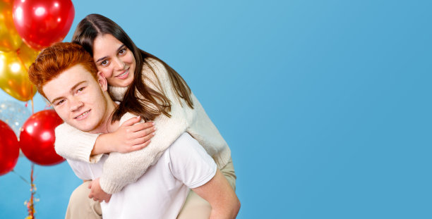 两个青少年在开玩笑，一个年轻漂亮的女孩坐在她的红发男友的背后，抓住他的脖子，气球在他们附近。图片下载