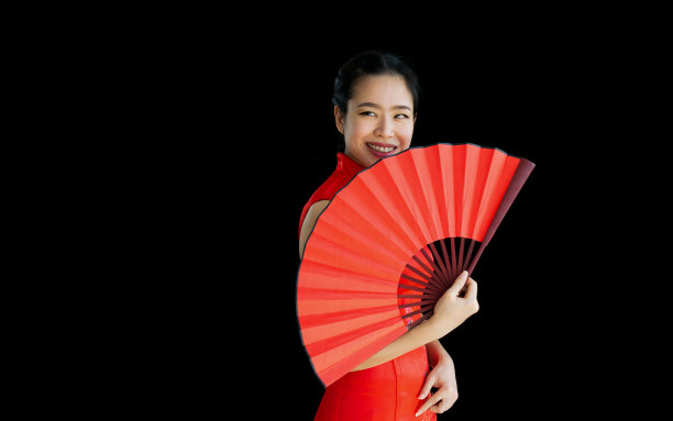 年轻的中国美丽的亚洲妇女穿着红色的传统服装，拿着折扇，站在黑色孤立的背景剪纸，看着相机，微笑着庆祝新年与拷贝空间。图片下载