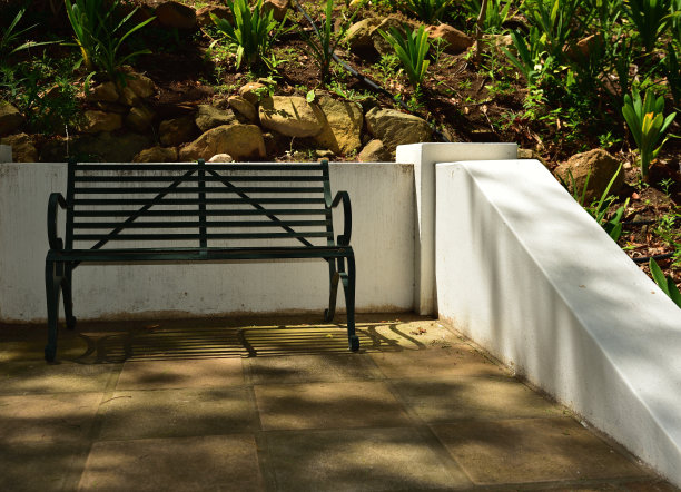 绿色的公园长椅靠在白色的墙壁上，周围是郁郁葱葱的植物，看起来很适合休息图片下载