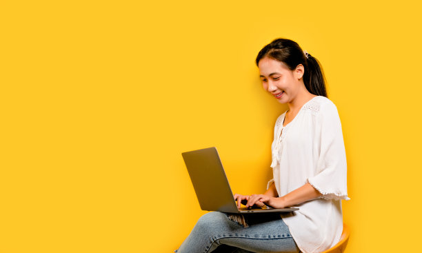 一个亚洲女人在他的笔记本电脑上工作，微笑着，在工作室的黄色背景上快乐的工作理念图片下载