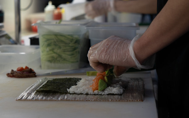这张照片是用米饭、黄瓜、红鱼、海藻和鳄梨制作寿司和卷的过程。男厨师手戴手套图片下载