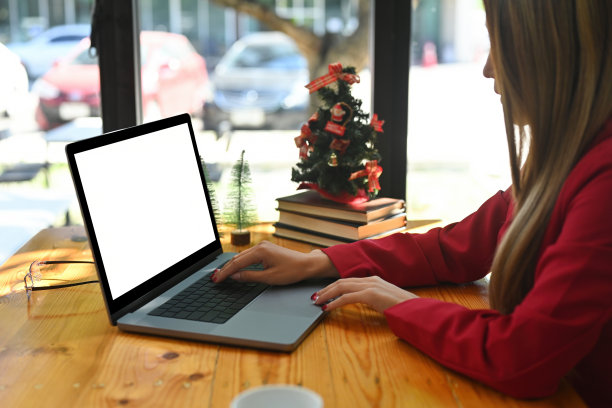 这是一张年轻的创意女性在木桌上用笔记本电脑打字的照片，木桌周围有咖啡杯、眼镜、一堆书和圣诞树。图片下载